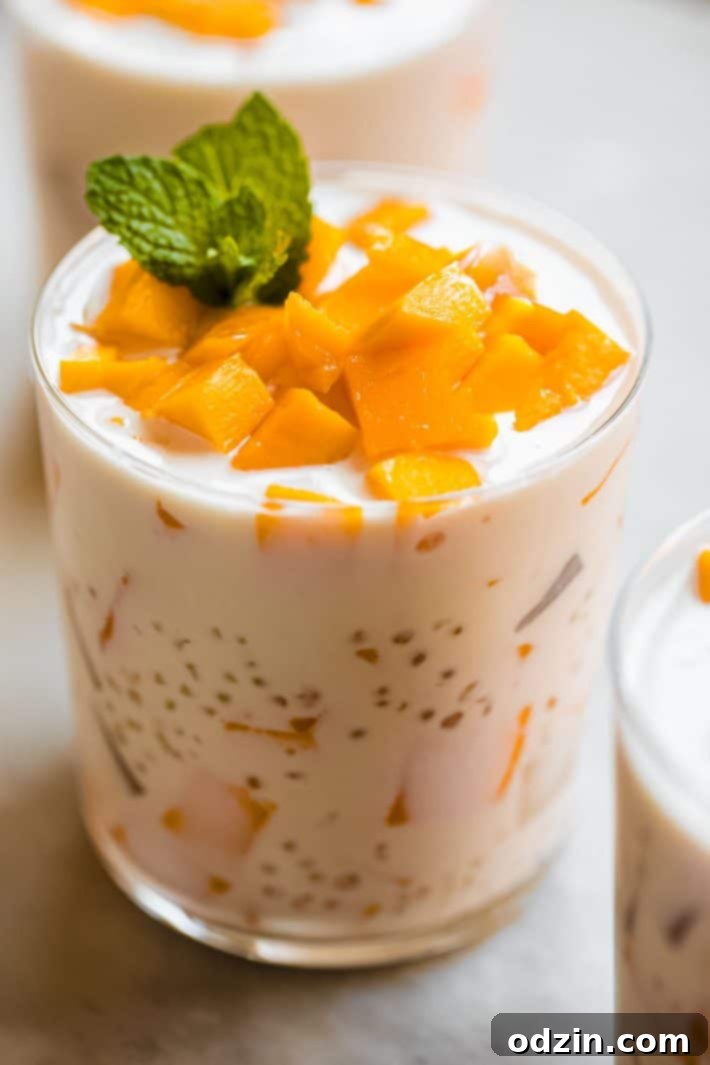 A close-up shot of a cup of Mango Bango dessert, showing layers of creamy pudding, tapioca pearls, and chunks of fresh mango, topped with a mint sprig.