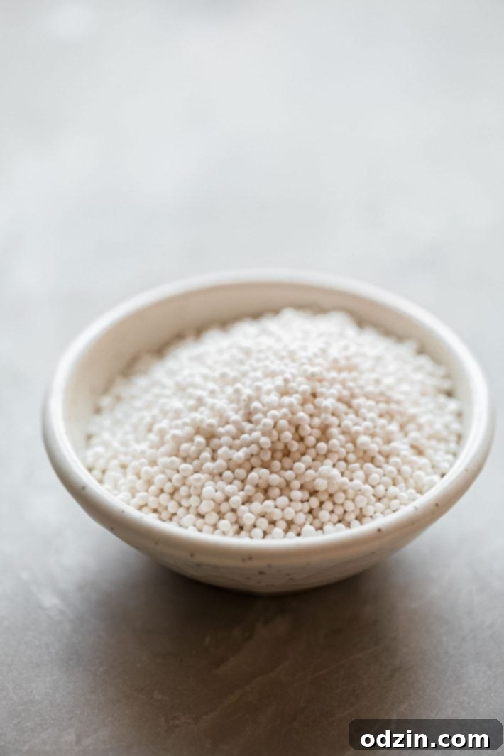 Small, translucent tapioca pearls resting in a speckled white bowl, ready for cooking, set on a clean white marble surface.