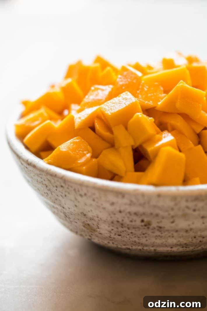 Freshly chopped, bright yellow mango chunks neatly arranged in a speckled white bowl, highlighting their juicy texture, on a white marble background.