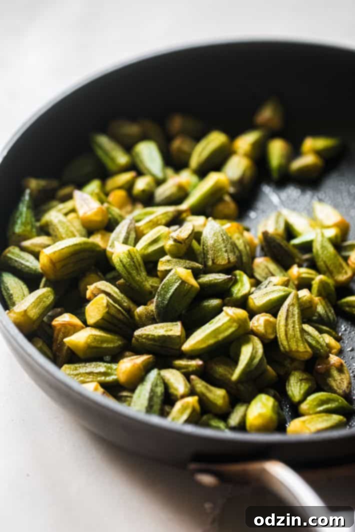 Fresh okra being sautéed in a frying pan with olive oil until lightly golden, preventing sliminess in the stew.