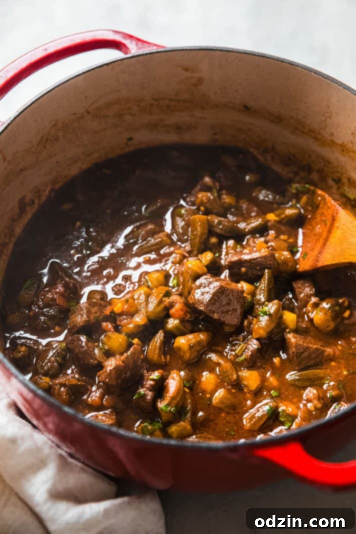 A large Dutch oven filled with bubbling Bamya (Middle Eastern Okra Stew with Beef), with a wooden spoon resting inside, ready to be served.