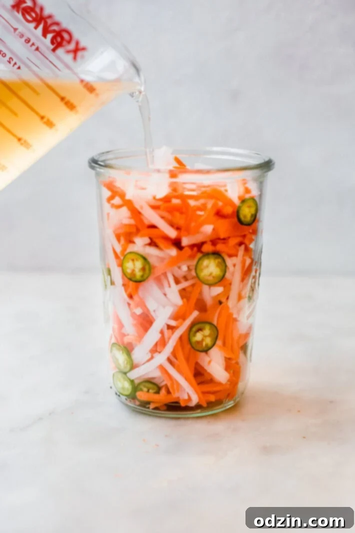Sliced daikon radish and carrots submerged in pickling liquid inside a glass jar, ready for the Banh Mi.