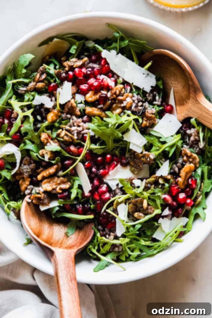 Vibrant holiday salad in a white serving bowl with elegant serving spoons, featuring arugula, ruby red pomegranate arils, nutty wild rice, and glistening candied nuts and seeds
