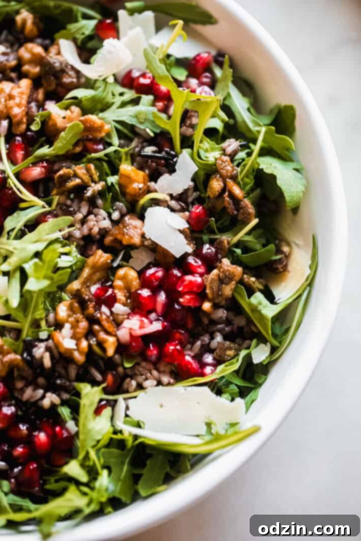 Close-up shot of the vibrant holiday salad, highlighting the texture of wild rice, the ruby red pomegranate seeds, fresh arugula leaves, and delicate cheese shavings, all ready to be dressed